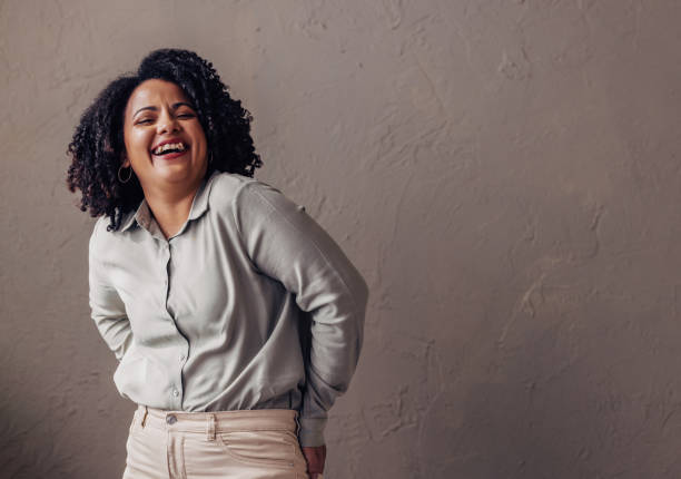 Joyful Woman Laughing Candidly in Stylish Attire A joyful woman with curly hair laughs candidly, dressed in a stylish light green blouse and beige trousers. Her genuine happiness is captured against a textured grey background. plus size woman fashion laughing stock pictures, royalty-free photos & images