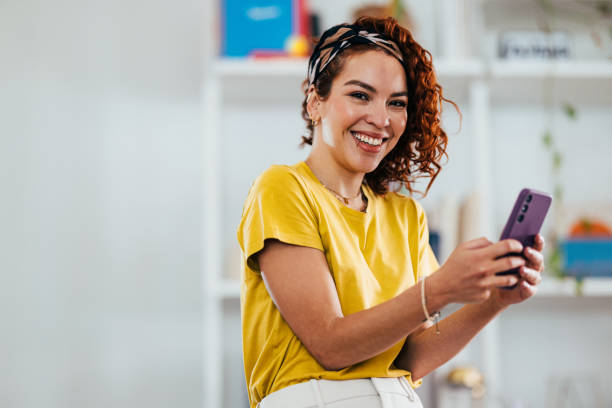 istock Smiling Woman in Yellow Top Using Smartphone Indoors 2151453402