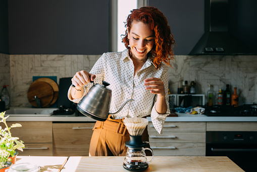 A cheerful woman with red curly hair pours hot water for coffee in a modern kitchen, displaying a joyful expression and casual style.