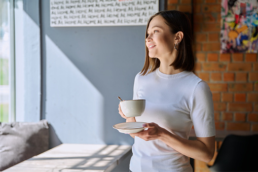 Young beautiful happy woman holding cup of coffee tea with saucer looking out window, gray wall of cafe coffee shop cafeteria copy space for text. Coffee business work services youth lifestyle leisure