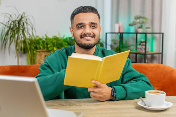 istock Indian man reading interesting book turning pages smiling enjoying literature taking a rest at table 2151484346