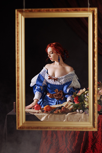 A woman in a blue medieval dress holds a pomegranate. The image has a vintage feel. Portrait of a woman in a Renaissance style dress with a still life.