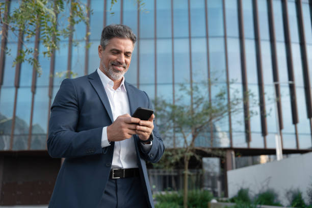 istock Smiling mature hispanic senior business man using smartphone cellphone at office building. Latin middle age male entrepreneur businessman holding mobile cell phone for trading outdoors. Copy space 2151530239