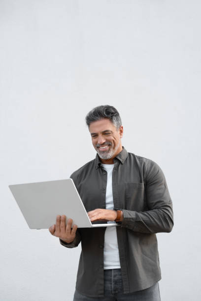 istock Mature smiling man using wireless pc for business solutions, isolated on white background. Middle age freelance entrepreneur, latin hispanic businessman holding laptop computer, working busy. Vertical 2151530251