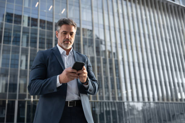 istock Mature hispanic senior business man using smartphone cellphone at office building. Focused serious latin middle age male entrepreneur businessman holding mobile cell phone for trading. Copy space 2151530261