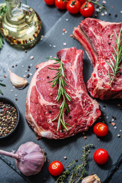 istock Two raw beef steaks on the bone on a black slate serving plate, decorated with various ingredients. 2151530328