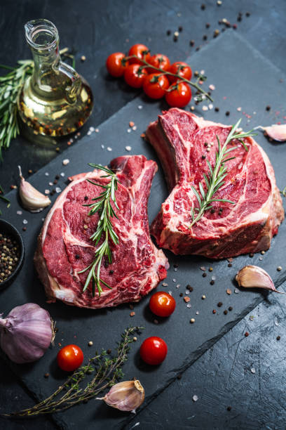 istock Two raw beef steaks on the bone on a black slate serving plate, decorated with various ingredients. 2151530374