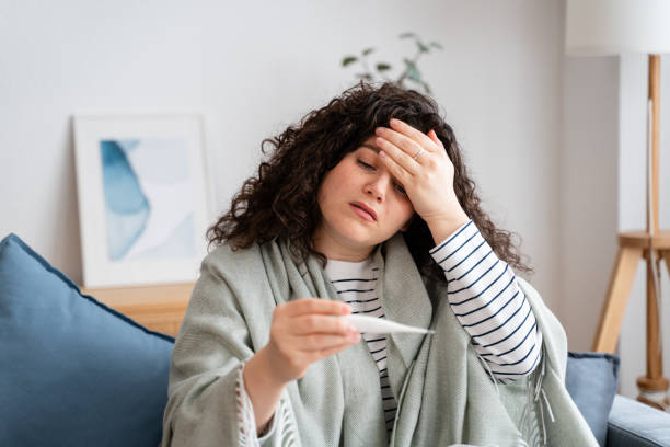 Unwell woman checking temperature at home Young woman feeling sick, checking her fever with a thermometer while sitting on a couch, looking exhausted and unwell fever stock pictures, royalty-free photos & images