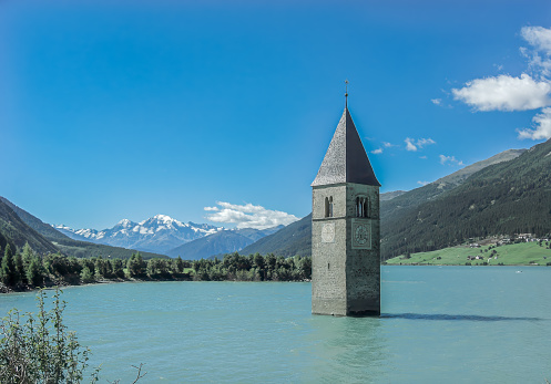 A picturesque tower sits in the middle of a tranquil lake, surrounded by majestic mountains under a sky filled with fluffy clouds. A stunning natural landscape perfect for travel enthusiasts