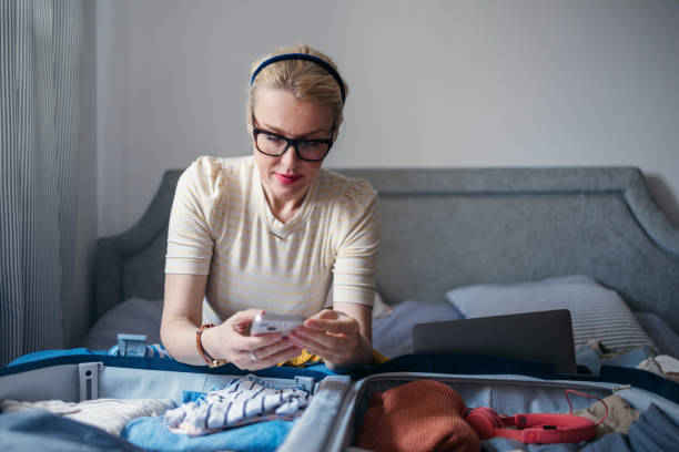 istock Serious Woman Using A Mobile Phone While Packing A Suitcase In The Bedroom 2151633230