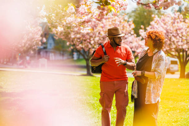 Multiracial mature couple enjoys a date and a walk under the cherry blossoms in James Park, Madison in New Jersey. African American bearded man in Fedora hat, gestures, and talks to his wife — Caucasian ginger haired woman amidst the trees. Focus on a family on background