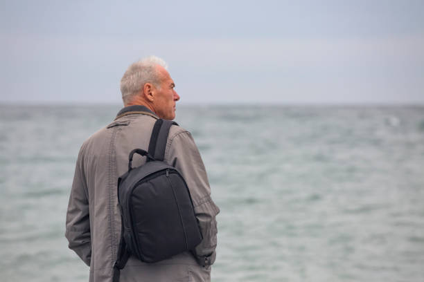 istock A gray-haired man with a backpack on his shoulder stands on the seashore. Close-up 2151642723