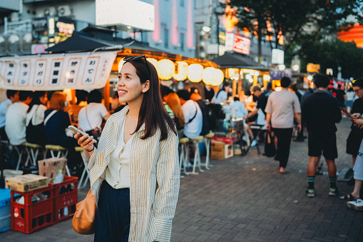 An Asian woman stands amidst a bustling night market, her gaze fixated on her phone. The festive mood is enhanced by the hanging lanterns and decorations, while chatter and bargaining create a lively atmosphere filled with joy and energy.