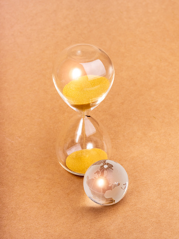 Environmental sustainability, time and responsibility, conserve resources, save planet concept. Small glass world globe near hourglass isolated on minimal brown paper background, vertical style.