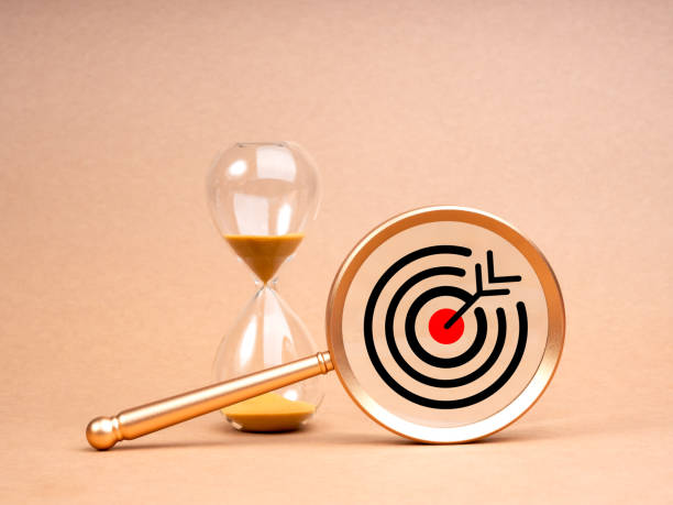 istock Short-term business goals encompass work that helps an organization reach its mid-term goals concept. Big target icon in gold magnifying glass lens near hourglass isolated on beige background. 2151651233