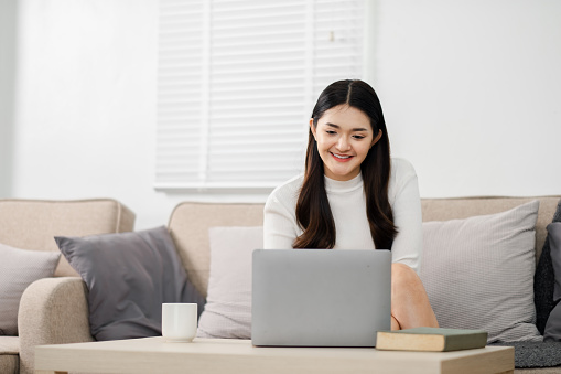 Young woman appears joyful as she uses her laptop while comfortably seated on a sofa in a well-lit living room.