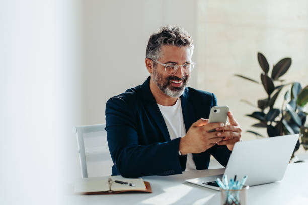 Focused professional man smiling at smartphone in bright modern office Cheerful, mature professional man is engrossed in his smartphone while sitting at a white desk in a sunny, contemporary office space. His laptop and notepad are at hand, suggesting productivity and connectivity. guy looking at cell phone stock pictures, royalty-free photos & images