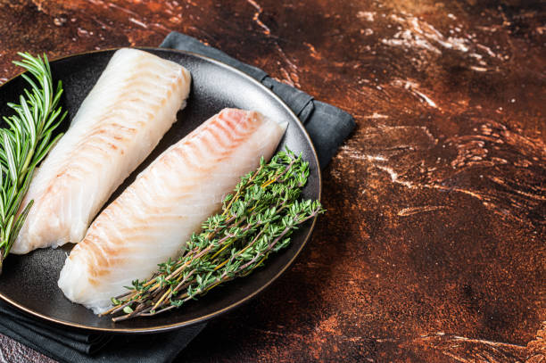 istock Raw cod loin fillet steaks with herbs on plate. Dark background. Top view. Copy space 2151678519