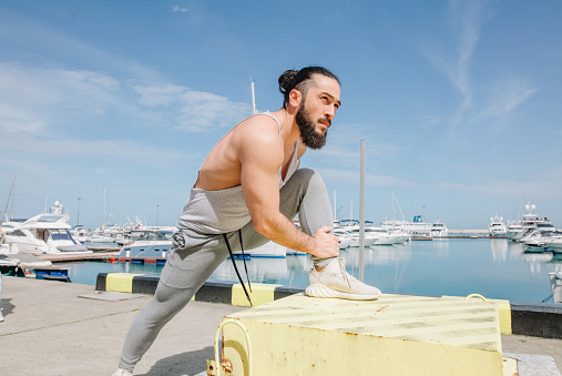 Athletic bearded sportsman with trendy hairstyle stretching legs while standing at the sea pier with sailing boats on background