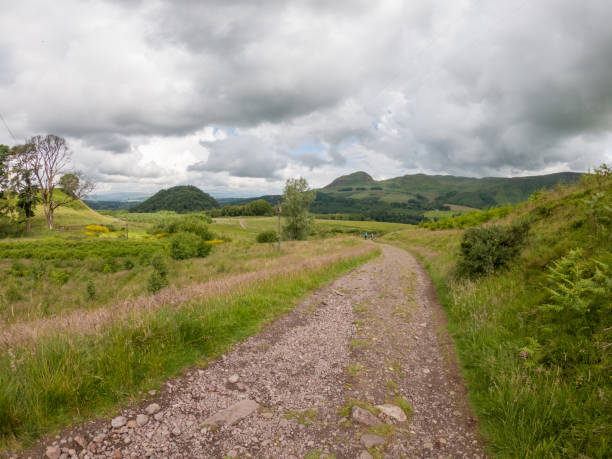 A gravel trail leading through lush meadows under a dramatic cloudy sky. Surrounded by rolling hills and scattered trees, the path invites exploration into the tranquil countryside. Distant figures can be seen enjoying the scenic walk, adding a sense of scale and activity to the landscape. The overcast sky adds a moody atmosphere to the vibrant greens and yellows of the natural setting, perfect for a peaceful hike.