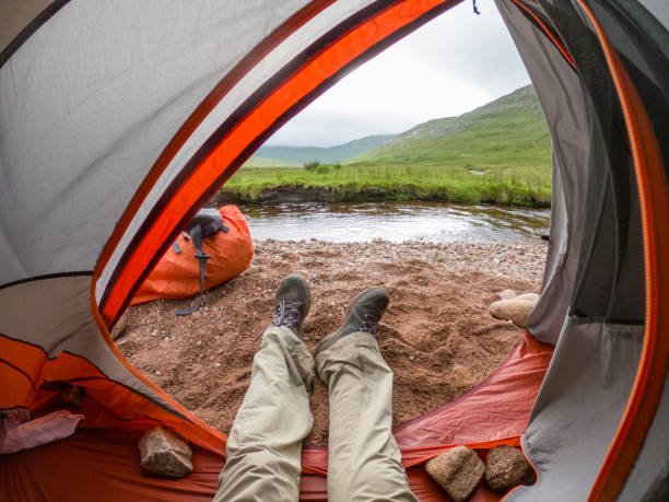 A camper's perspective from inside a tent, overlooking a tranquil stream set against the lush greenery of the Scottish Highlands. The view focuses on the camper's legs stretched out, with hiking boots on, showing a relaxed pose within the tent. The open tent flap frames a picturesque scene of rolling hills and a gentle stream, promoting a sense of peace and the perfect retreat into nature.