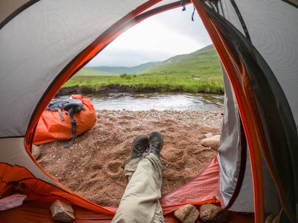 A camper's peaceful morning view from within a tent, situated by a gentle highland stream. The perspective centers on the camper's legs extending from the tent onto the sandy riverbank, emphasizing a relaxed atmosphere. The tent's open flap reveals a stunning vista of lush green hills and the meandering stream, offering a picturesque escape into the wilderness.