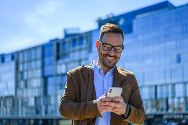 istock Smiling businessman texting over smart phone while standing against modern office building in city 2151691339