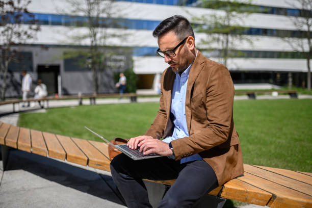 istock Serious entrepreneur in glasses working on project over laptop while sitting on bench in modern city 2151691499