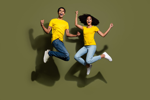 Full size photo of two young people jump raise fists wear t-shirt isolated on khaki color background.