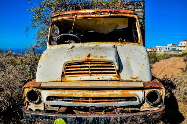An old, rusted truck is parked in a field. The truck is covered in dirt and has a faded white paint job. The front of the truck has a large dent in the hood, real image