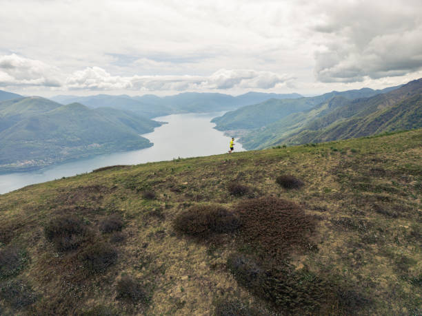 A solitary runner in motion along a rugged mountain trail, with a vast and scenic lake stretching into the distance surrounded by lush green mountains. Dressed in a bright yellow top, the runner's energetic stride and focus add a vivid contrast to the soft, overcast sky and the natural tranquility of the expansive landscape.