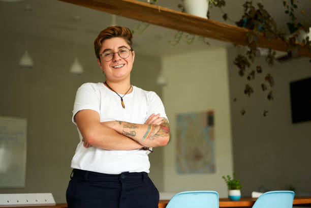 Transgender young adult smiles, arms crossed, wearing casual attire, representing workplace inclusion, gender identity pride. Confident Gen Z individual with tattoos stands in modern office. individuo seguro de la generación z con tatuajes se encuentra en una oficina moderna. joven adulto transgénero sonríe, con los brazos cruzados, viste atuendo informal, representa la inclusión en el lugar de trabajo, el orgullo de identidad de género.