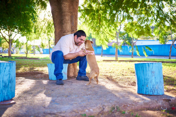 Portrait of a Latin man playing with his dog, in a park, sitting under a big tree.