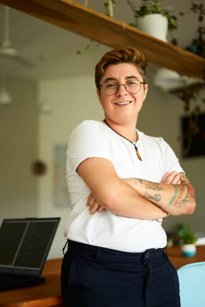 istock Confident transgender pro in office attire stands with arms folded, smiling at camera. Trans exec in well-lit workspace with laptop. Genderqueer identity embraced. 2151720672