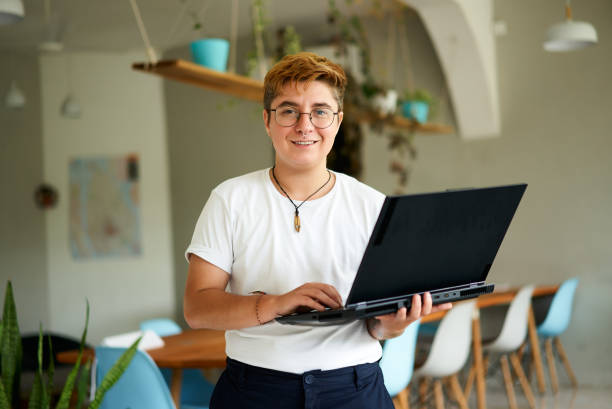 FTM professional in casual attire stands, ready for business tasks. Confident transgender office worker with laptop in modern workspace. Inclusive workplace culture, gender diversity reflected. oficinista transgénero segura de sí misma con computadora portátil en un espacio de trabajo moderno. ftm profesional en stands de atuendo casual, listo para tareas comerciales. cultura inclusiva en el lugar de trabajo, diversidad de género reflejada.