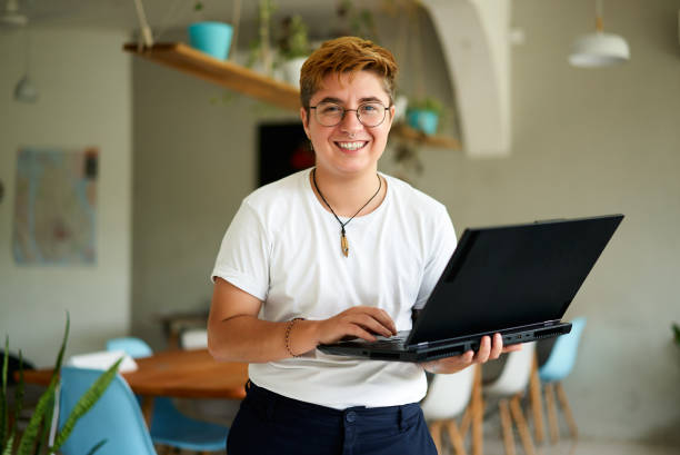 Cheerful transgender professional holds laptop, smiles in modern office. Gen Z using tech for work, represents inclusivity. Proud, confident posture, casual style, indoor plants background.