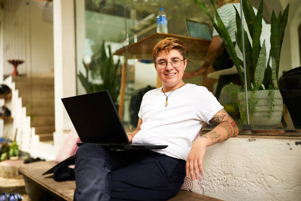 Inked, digital nomad in casual clothes, emphasizing diversity, inclusivity in modern remote work settings. Transgender Gen Z freelancer smiles while working on laptop at eco-friendly coworking space. un autónomo transgénero de la generación z sonríe mientras trabaja en un portátil en un espacio de coworking ecológico. entintado, nómada digital con ropa informal, enfatizando la diversidad y la inclusión en entornos modernos de trabajo remoto.