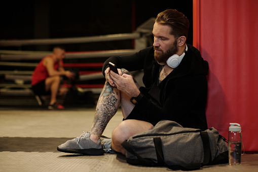 Young serious male boxer with tattoos on leg and headphones on neck sitting on the floor in gym after training and texting in mobile phone