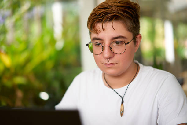 istock Transgender pro focuses laptop work in office. Inclusive environment, modern workplace diversity. Confident individual with glasses, casual attire. Background plants indicate eco-friendly space. 2151726629