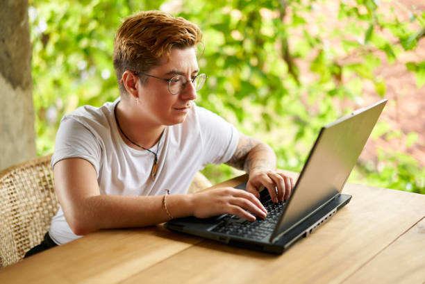 istock Transgender digital nomad types on laptop at tropical coworking space. Non-binary entrepreneur focuses on remote work among green plants. Inclusive, modern telecommuting lifestyle in natural setting. 2151726976