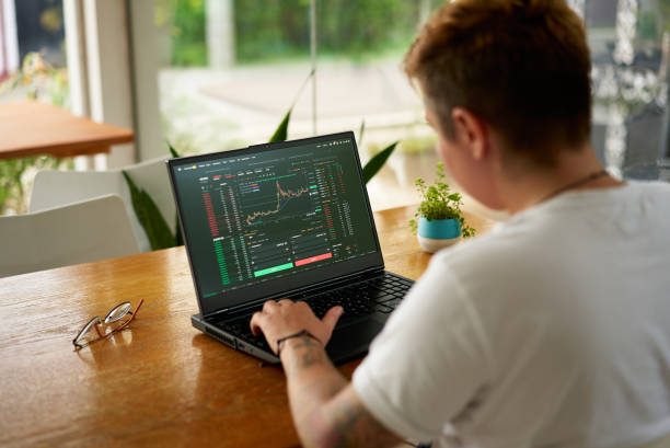 istock Pro at work desk studies financial markets on computer, analyzing live trading charts. Focused on investment trends. Home office setup, casual attire, daytime, indoor financial activity. 2151728664