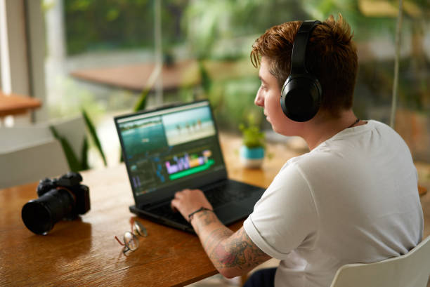 istock Focused individual with headphones edits on laptop inside cafe, video editing skills at work. mirrorless camera lays beside, freelance job in progress. Cafe surroundings, daylight illuminates process. 2151728821