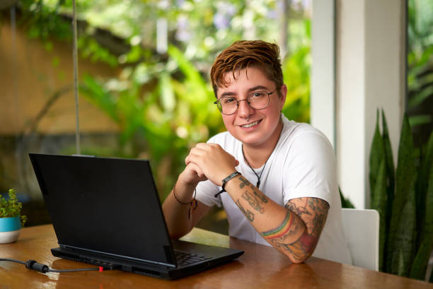 istock Smiling transgender man works on laptop in modern coworking space. Comfortable inclusive work environment, happy LGBT pro using tech, earphones on desk, indoor plants, natural light. 2151729119