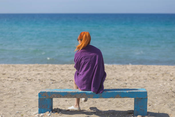 Woman cloaked with towel sitting on bench on sand beach