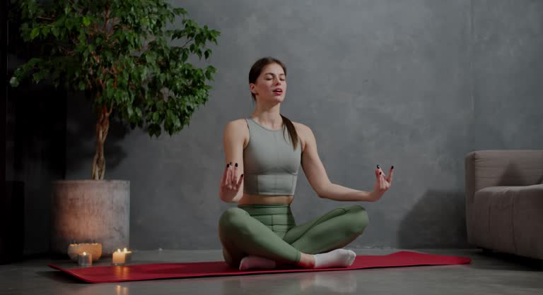 Confident and calm brunette girl in a gray top and green sweatpants sits on a red sports carpet and does yoga near lit candles and a home houseplant in a modern apartment