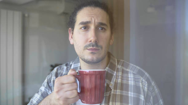 Depressed young male looks through window while drinking tea