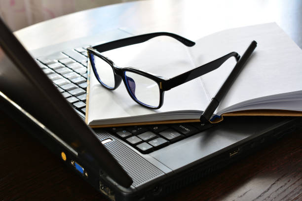 Pile of unfinished paperwork on office desk beside notebook with bar graph on screen. Stack of homework assignment with plastic slide binding bas. Report papers stacks. Business and education concept.