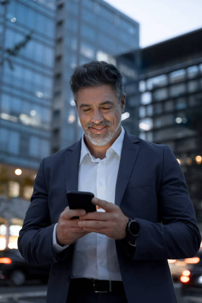 istock Smiling mature hispanic senior business man using app, holding smartphone cellphone at office building. Indian or latin middle age male entrepreneur businessman working on mobile cell phone. Vertical 2151777346