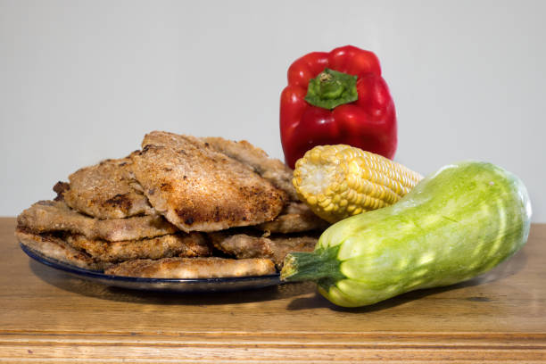 stack of breaded milanesas on a blue-rimmed plate placed on a wooden table next to a corn, a red pepper and a zuchini squash. The background is out of focus. Milanesas appear to be baked