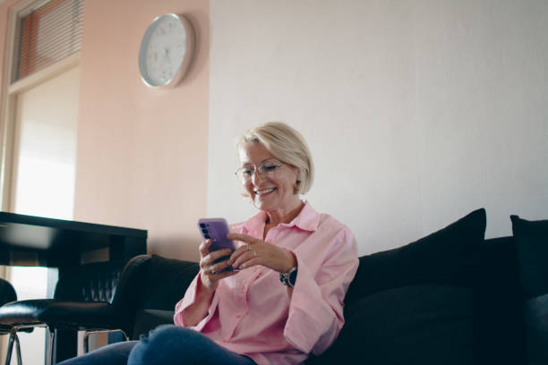 istock mature blond woman sitting on sofa and using mobile 2151780785